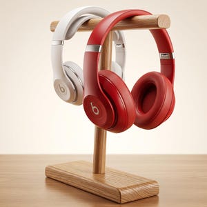 Op de afbeelding: Een paar over-ear hoofdtelefoons, één wit en één rood, beide voorzien van het Beats-logo op de oorschelpen, worden gepresenteerd op een lichthouten standaard. De standaard bestaat uit een rechthoekige voet en een paal met een dwarsbalk.