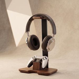 Puede incluir: Un soporte para auriculares de madera oscura y metal negro con una barra superior horizontal de madera para colgar auriculares y una base inferior de madera para un mando de videojuegos blanco y negro sobre una base rectangular.