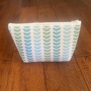 May include: A zippered pouch featuring a repeating vertical pattern of stylized leaves in alternating shades of light blue and sage green against a white background. The rectangular bag has a white zipper closure along the top edge.