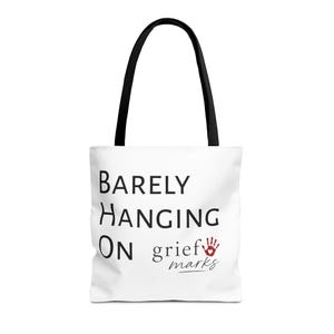 May include: A white tote bag with black handles featuring the black text BARELY HANGING ON and the text grief marks in black script with a red handprint icon containing a small white heart shape centred on the front of the bag.