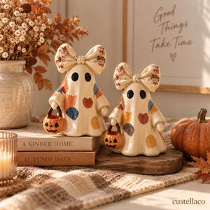 Peut inclure: Deux figurines de fantômes couleur crème avec de grands nœuds sur la tête, ornées de motifs en patchwork colorés et de détails floraux. Chaque fantôme a des yeux ovales noirs et tient un petit seau citrouille orange à la main.