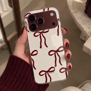 May include: A clear smartphone case featuring a repeating pattern of dark red bows. The case has a dark red camera module cutout area at the top. The design is printed on a transparent background, allowing the phone's surface to remain visible underneath.