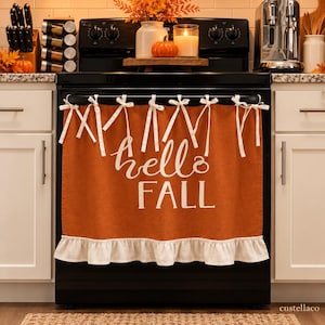 Peut inclure: Un cache-four orange brûlé avec le texte blanc "hello FALL" écrit en lettres cursives et capitales. Le panneau en tissu est fixé à la poignée du four par des rubans blancs et présente un ourlet à volants blanc sur le bord inférieur.