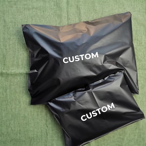 Puede incluir: Dos sobres de envío rectangulares de plástico negro de diferentes tamaños, cada uno con la palabra CUSTOM impresa en letras mayúsculas blancas y negritas centradas en la parte frontal. Las bolsas tienen un acabado liso y reflectante.