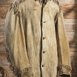 May include: A tan suede leather jacket featuring long fringe detailing along the shoulders and sleeves. The jacket has a button-down front with metallic buttons and embroidered floral patterns on the cuffs of the long sleeves.