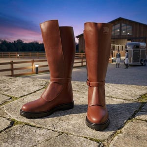 May include: A pair of knee-high brown leather riding boots featuring a smooth finish, a rounded toe, a thick black lug sole, and a decorative strap detail around the ankle. The boots have a structured shaft with a subtle embossed logo near the top.