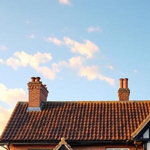 May include: A brick house exterior featuring a dark brown tiled roof with two prominent brick chimneys. The structure includes a white-framed window and dark timber framing accents against the red brick wall under a clear blue sky.