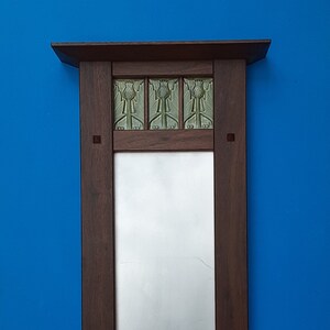 Puede incluir: Un espejo de pared rectangular con un marco de madera marr&oacute;n oscuro que presenta un peque&ntilde;o estante en la parte superior. Tres azulejos de cer&aacute;mica verdes cuadrados con un motivo de cardo en relieve est&aacute;n incrustados sobre el espejo.