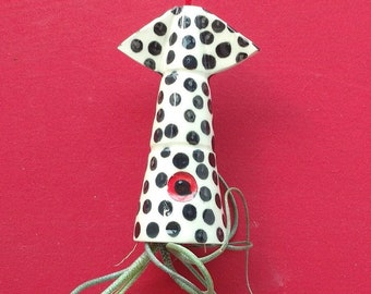 May include: A white ceramic planter with black polka dots and a red circle in the center. The planter is hanging from a red string and has a green air plant growing out of it.