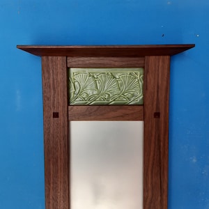 May include: A wooden mirror with a decorative green tile inset featuring a stylized ginkgo leaf design. The mirror is framed in dark wood with a simple, rectangular shape.