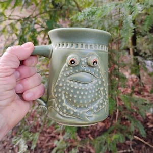 Puede incluir: Una taza de cerámica verde con un diseño de cara de rana. La taza tiene un asa y una cara de rana detallada con ojos saltones y una boca sonriente. La taza es de color verde claro y tiene una superficie texturizada.