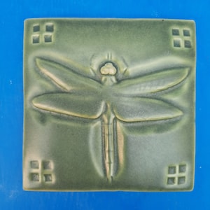May include: A square sage green ceramic tile featuring a raised relief design of a dragonfly in the center. The four corners of the tile each contain a small square motif consisting of four smaller recessed squares arranged in a grid pattern.