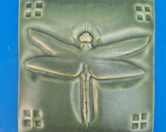 May include: A green ceramic tile with a raised dragonfly design. The dragonfly has a light green body and wings. The tile has a square shape with a border of four small squares.