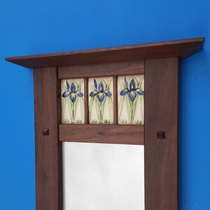 K&ouml;nnte beinhalten: Ein rechteckiger Holzspiegelrahmen mit dunkelbraunem Finish und einer oberen Ablage. Der obere Bereich enth&auml;lt drei quadratische Keramikfliesen, die jeweils ein handgemaltes Motiv einer blauen und gelben Irisbl&uuml;te auf cremefarbenem Grund zeigen.