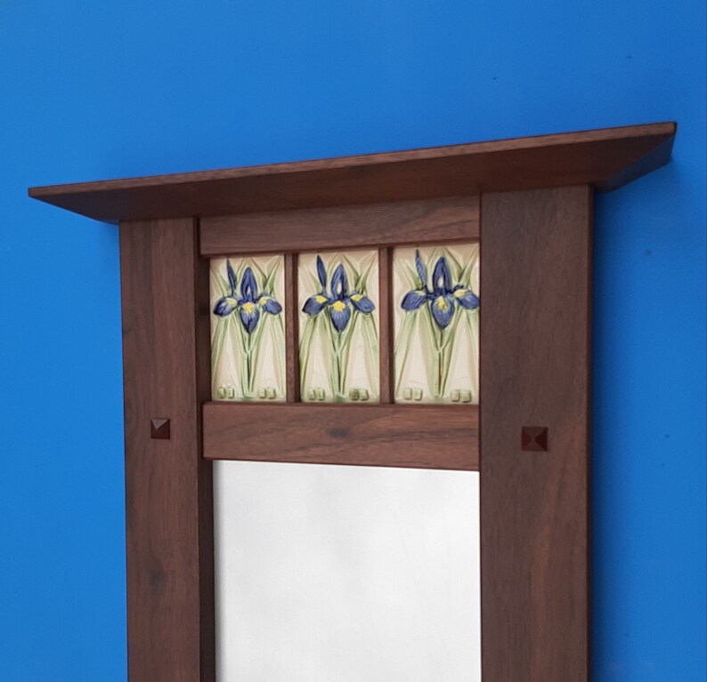 K&ouml;nnte beinhalten: Ein rechteckiger Holzspiegelrahmen mit dunkelbraunem Finish und einer oberen Ablage. Der obere Bereich enth&auml;lt drei quadratische Keramikfliesen, die jeweils ein handgemaltes Motiv einer blauen und gelben Irisbl&uuml;te auf cremefarbenem Grund zeigen.