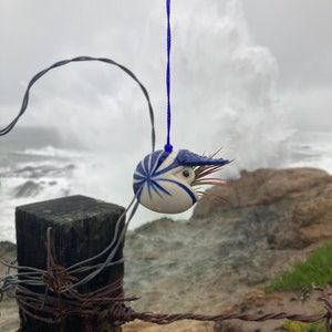 May include: A blue and white ceramic nautilus shell pendant with a red and white feather hanging from it. The pendant is hanging from a blue string against a backdrop of a stormy ocean.
