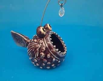 May include: A dark brown ceramic anglerfish figurine with a round, textured body, wide open mouth featuring small teeth, and two prominent eyes. A thin silver wire extends from the top, holding a small, clear, faceted crystal pendant.