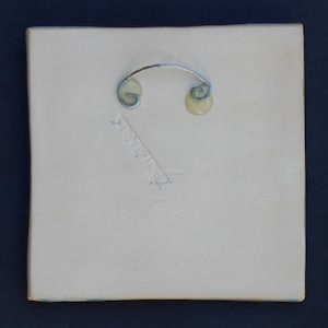 May include: A square, off-white ceramic tile featuring a silver-toned metal wire hanger attached to the back with two circular adhesive mounts. The back surface displays an impressed signature that reads CINDY ZIMMER in a diagonal orientation.