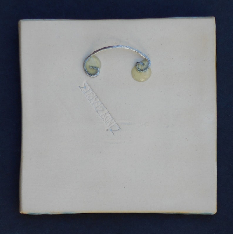 May include: A square, off-white ceramic tile featuring a silver-toned metal wire hanger attached to the back with two circular adhesive mounts. The back surface displays an impressed signature that reads CINDY ZIMMER in a diagonal orientation.