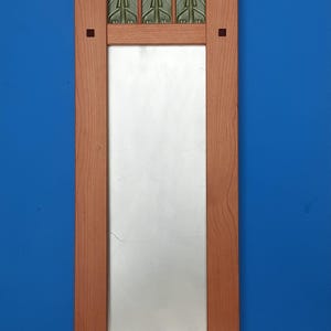 K&ouml;nnte beinhalten: Ein rechteckiger Wandspiegel mit einem hellbraunen Holzrahmen. Der Rahmen verf&uuml;gt &uuml;ber eine flache Ablage oben, vertikale Seitenteile mit kleinen quadratischen Einlagen und eine horizontale untere Leiste. Drei gr&uuml;ne Zierfliesen mit gepr&auml;gten Mustern befinden sich &uuml;ber dem Glas.