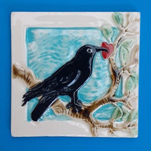 May include: Square ceramic tile featuring a black crow perched on a branch, holding a red heart in its beak. The background has a light blue and white design. The tile is framed with a white border. The crow and branch are brown and green.