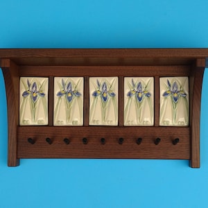 May include: A wooden wall-mounted key rack with a shelf and five ceramic tiles. Each tile features a blue and yellow iris flower design. The rack has seven black hooks for keys and a dark brown finish.