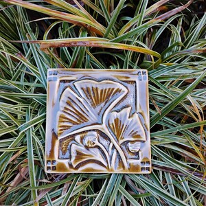 May include: A square ceramic tile featuring a glossy, dark brown glaze with a relief-molded ginkgo leaf design. The tile has a raised border with small square accents at each corner, showcasing a botanical motif with deep, textured grooves.