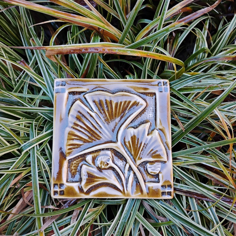 May include: A square ceramic tile featuring a glossy, dark brown glaze with a relief-molded ginkgo leaf design. The tile has a raised border with small square accents at each corner, showcasing a botanical motif with deep, textured grooves.