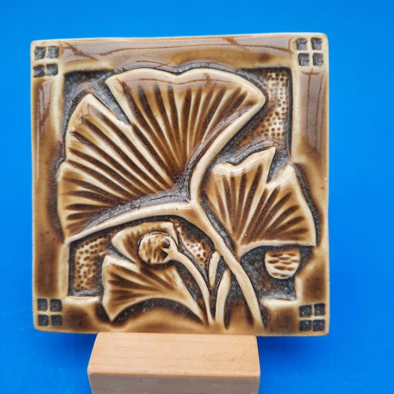 May include: A square ceramic tile featuring a relief design of ginkgo leaves in shades of brown and tan. The surface has a glossy finish with dark brown recessed areas and lighter brown raised details, framed by small square patterns in the corners.