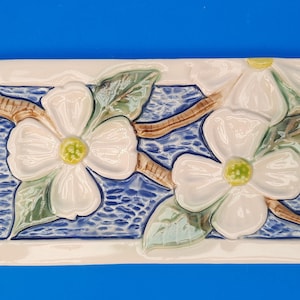 May include: A ceramic tile with a raised design of white dogwood blossoms with green leaves on a blue background.