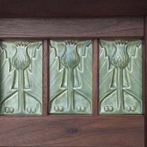 Puede incluir: Tres azulejos de cer&aacute;mica rectangulares de color verde salvia con un motivo de cardo en relieve, colocados dentro de un marco de madera marr&oacute;n oscuro. Cada azulejo muestra una flor de cardo central con hojas y el marco incluye detalles cuadrados.