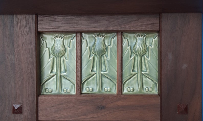 Puede incluir: Tres azulejos de cer&aacute;mica rectangulares de color verde salvia con un motivo de cardo en relieve, colocados dentro de un marco de madera marr&oacute;n oscuro. Cada azulejo muestra una flor de cardo central con hojas y el marco incluye detalles cuadrados.