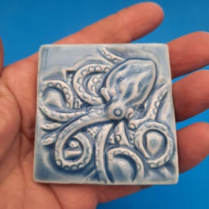 May include: A blue ceramic tile with a raised design of an octopus with its tentacles curled around it. The octopus is in a light blue glaze.