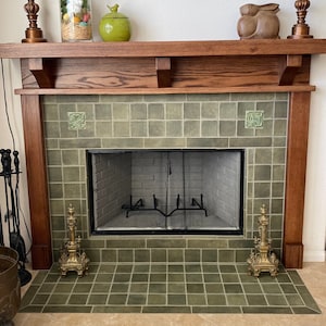 May include: A fireplace featuring a dark wood mantel, sage green square tiles, and a black metal mesh screen. Two ornate brass andirons sit on the hearth. Decorative items include a glass jar, a green ceramic jar, and a wooden rabbit sculpture.