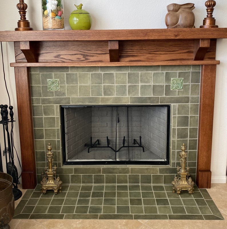 May include: A fireplace featuring a dark wood mantel, sage green square tiles, and a black metal mesh screen. Two ornate brass andirons sit on the hearth. Decorative items include a glass jar, a green ceramic jar, and a wooden rabbit sculpture.