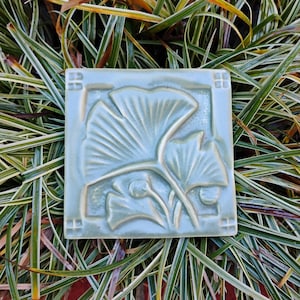 May include: A square ceramic tile in a soft sage green glaze featuring a raised relief design of two ginkgo leaves. The tile has a smooth, glossy finish with a border frame that includes small square accents at each of the four corners.