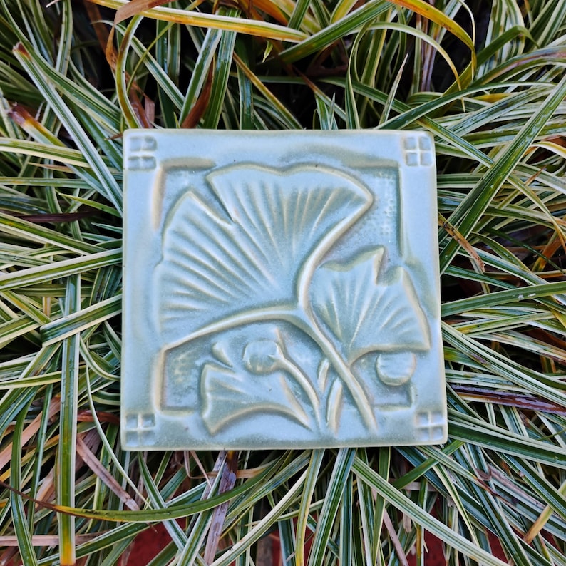 May include: A square ceramic tile in a soft sage green glaze featuring a raised relief design of two ginkgo leaves. The tile has a smooth, glossy finish with a border frame that includes small square accents at each of the four corners.