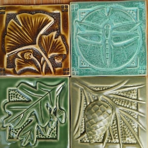 May include: Four square ceramic tiles featuring embossed nature designs. The top left tile is brown with ginkgo leaves. The top right is teal with a dragonfly. The bottom left is green with oak leaves and acorns. The bottom right is olive with a pinecone.