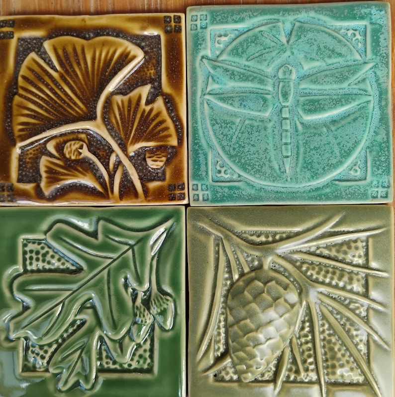 May include: Four square ceramic tiles featuring embossed nature designs. The top left tile is brown with ginkgo leaves. The top right is teal with a dragonfly. The bottom left is green with oak leaves and acorns. The bottom right is olive with a pinecone.