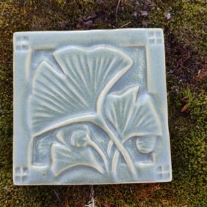 May include: A square ceramic tile with a glossy sage green glaze featuring a raised relief design of ginkgo leaves. The tile has a smooth surface with a border containing small square indentations at each corner, highlighting the botanical motif.