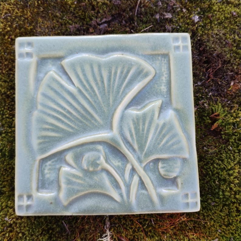 May include: A square ceramic tile with a glossy sage green glaze featuring a raised relief design of ginkgo leaves. The tile has a smooth surface with a border containing small square indentations at each corner, highlighting the botanical motif.