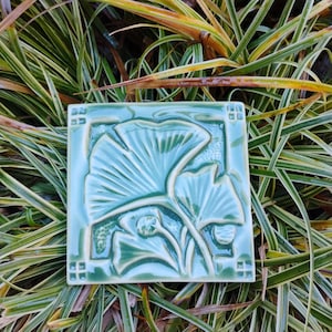 May include: A square ceramic tile in a glossy sage green glaze featuring a raised relief design of ginkgo leaves. The tile has a decorative border with small square accents in each corner, showcasing a smooth, reflective surface finish.