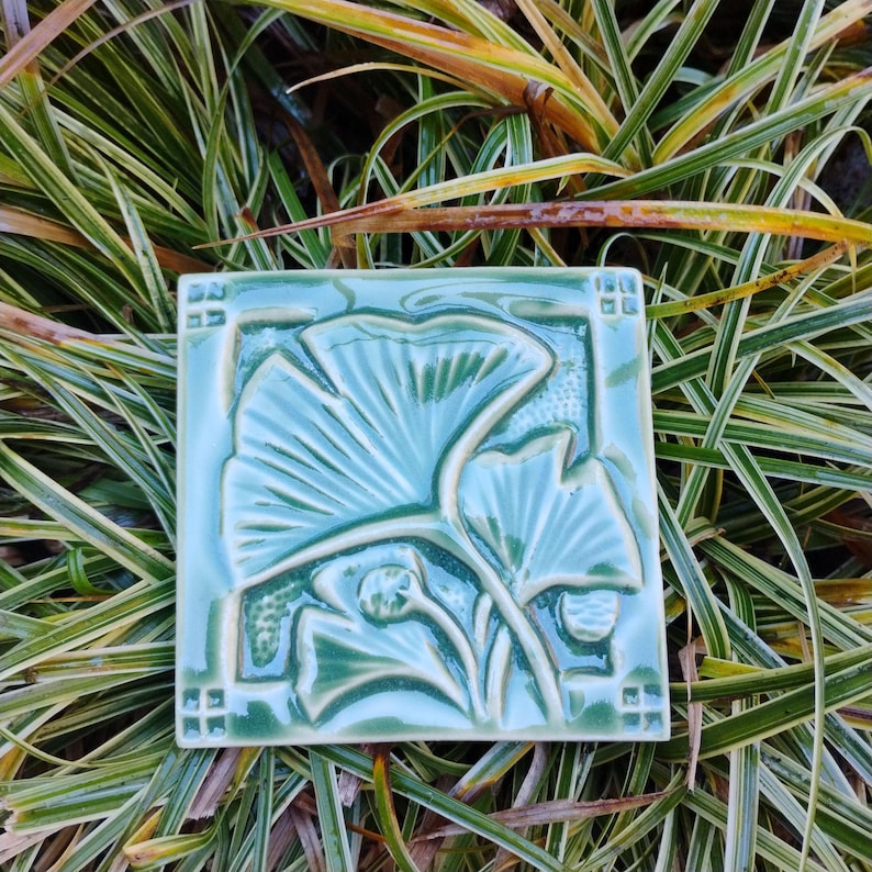 May include: A square ceramic tile in a glossy sage green glaze featuring a raised relief design of ginkgo leaves. The tile has a decorative border with small square accents in each corner, showcasing a smooth, reflective surface finish.
