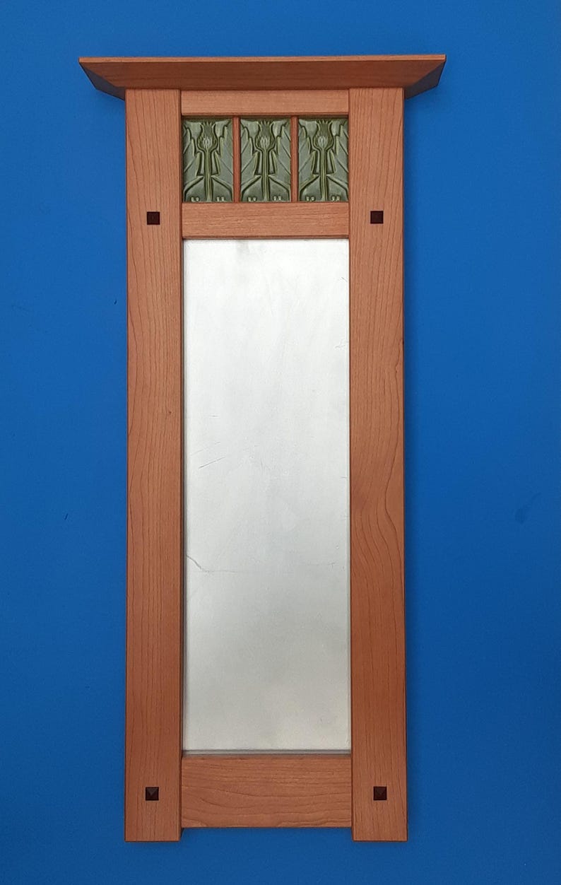 Puede incluir: Un espejo de pared rectangular con un marco de madera de color marr&oacute;n claro. El marco cuenta con un estante superior plano, paneles laterales verticales con peque&ntilde;as incrustaciones cuadradas y un riel inferior horizontal. Tres azulejos decorativos verdes con patrones en relieve se sit&uacute;an sobre el cristal.