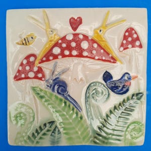May include: A ceramic tile with a whimsical woodland scene. A red and white mushroom with yellow birds perched on top, a blue bird, a snail, and green ferns. The tile has a white background.
