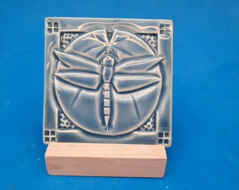 May include: A blue ceramic tile with a raised design of a dragonfly in a circle. The tile is on a wooden stand.