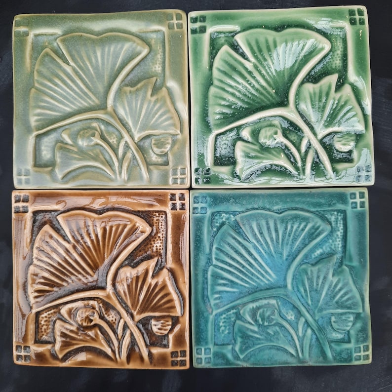 May include: Four square ceramic tiles featuring a raised relief design of ginkgo leaves. The tiles are glazed in shades of sage green, emerald green, warm brown, and teal blue, each with a decorative border of small squares in the corners.