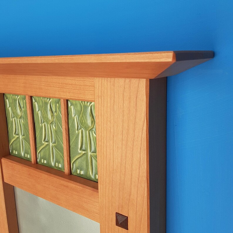 Puede incluir: Un marco de espejo de madera con acabado de veta de madera marr&oacute;n claro y un estante superior. La secci&oacute;n superior contiene tres azulejos de cer&aacute;mica verde claro, cada uno con un dise&ntilde;o floral estilizado en relieve. Se observa una peque&ntilde;a incrustaci&oacute;n cuadrada oscura.