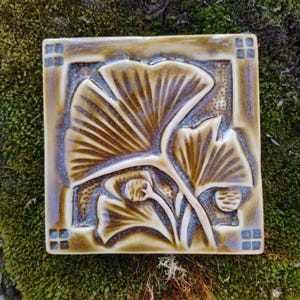 May include: A square ceramic tile featuring a relief design of ginkgo leaves in shades of brown and tan. The tile has a glossy finish with a dark border and small square patterns in each corner, showcasing a detailed botanical motif.