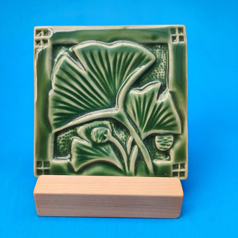 May include: A square ceramic tile featuring a raised relief design of ginkgo leaves in varying shades of deep green. The tile has a glossy finish and is displayed on a rectangular light-colored wooden stand, showcasing intricate leaf veins.